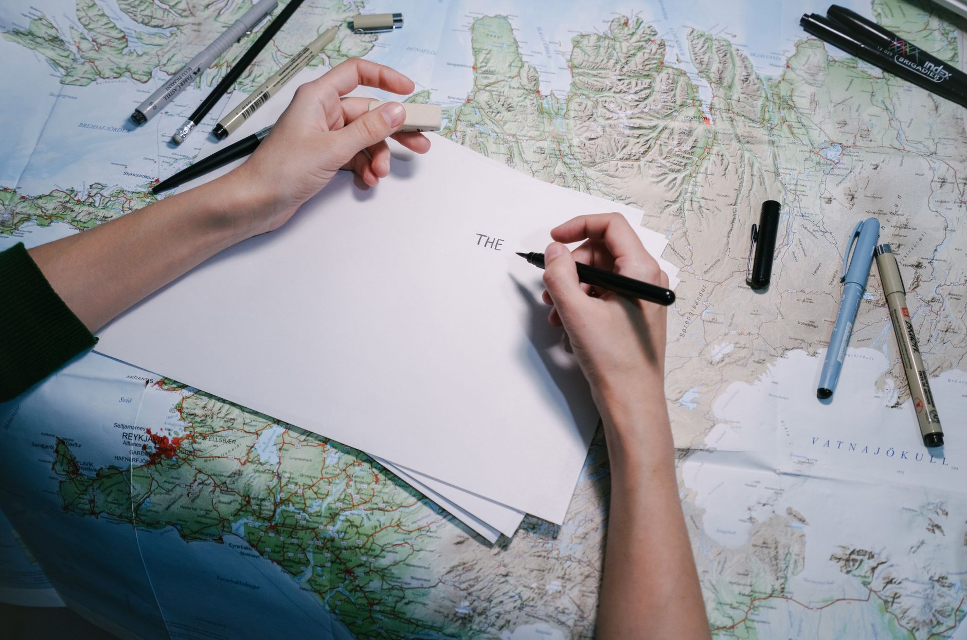 Hands writing on paper over a detailed world map with various pens.