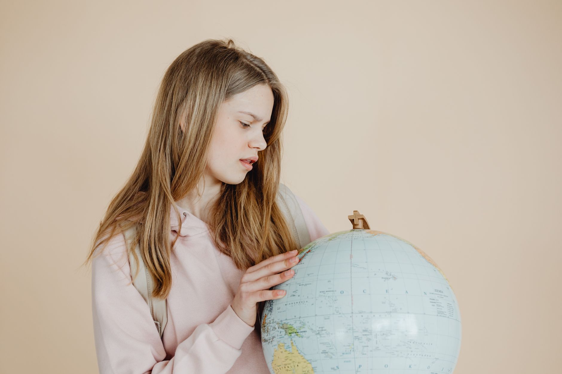 Teen girl in pink hoodie studying a globe against a neutral background. Education concept.