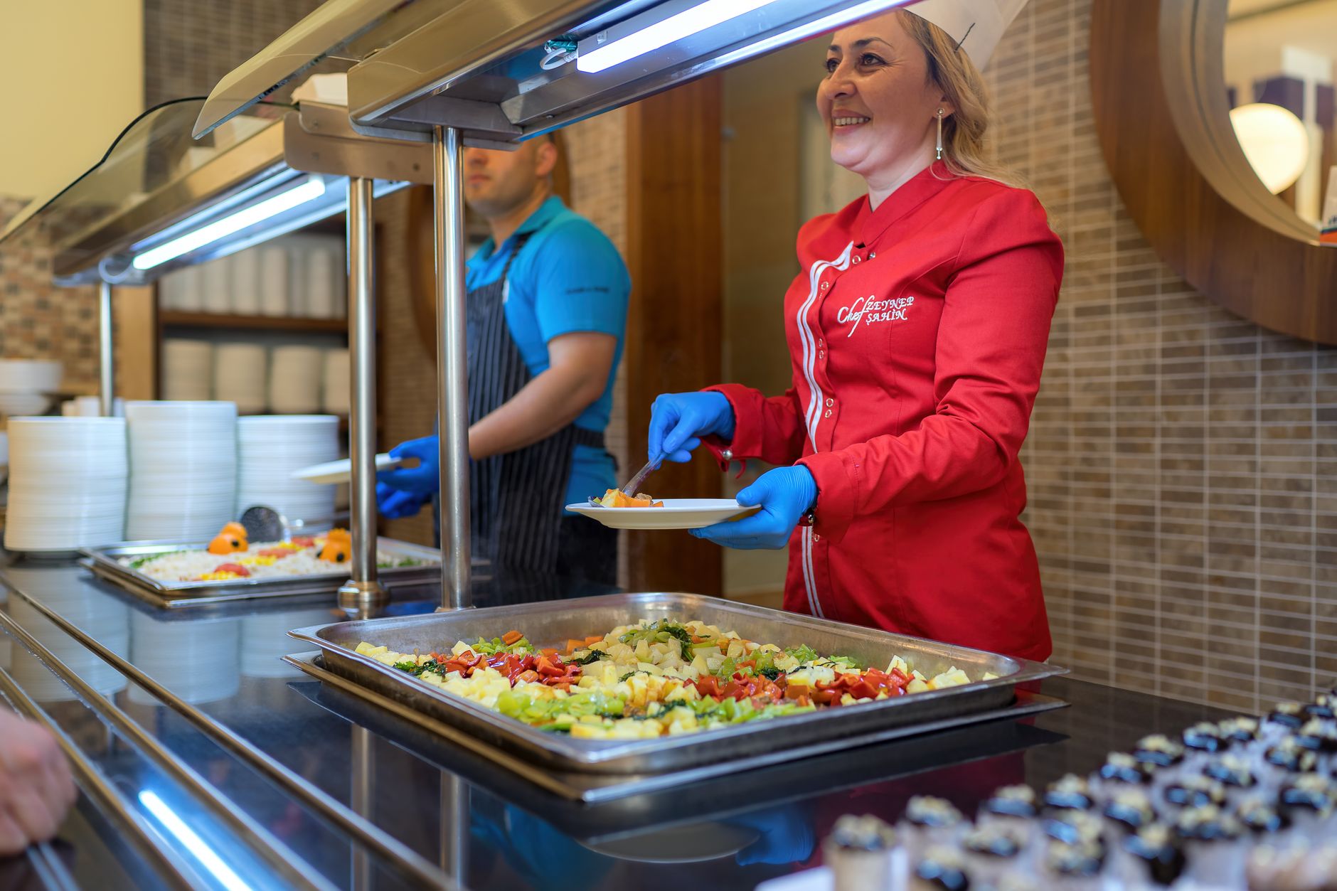 Chef in red uniform serving food at a buffet counter in Antalya restaurant, smiling and engaging with customers.