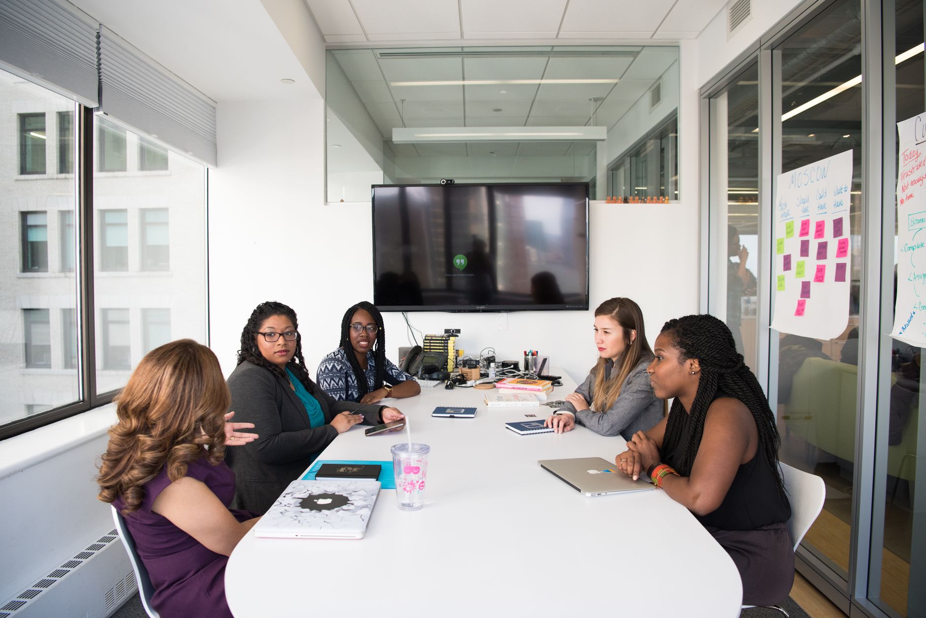 Five diverse women collaborating in a modern office meeting room, discussing projects.