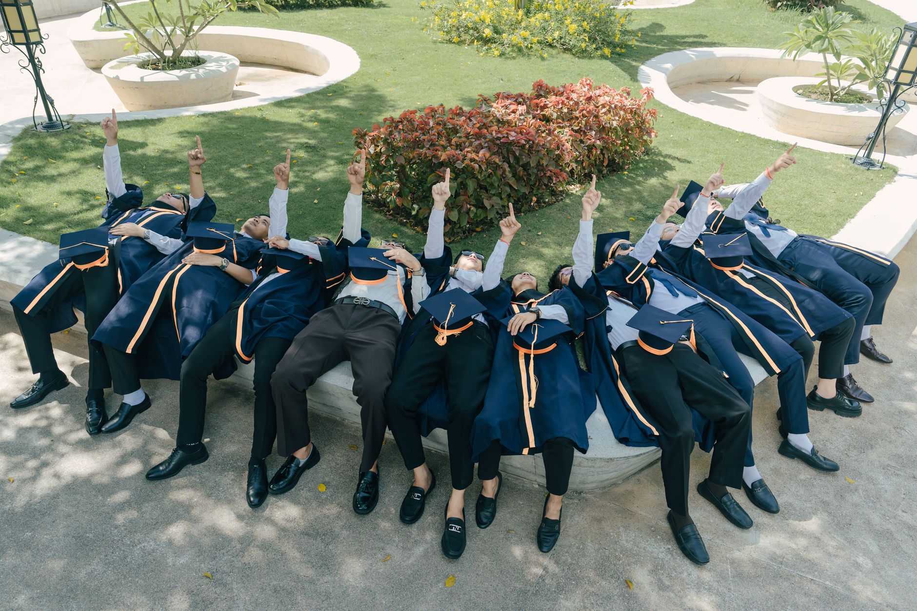 Graduates in caps and gowns celebrate by pointing skyward outdoors on a sunny day.