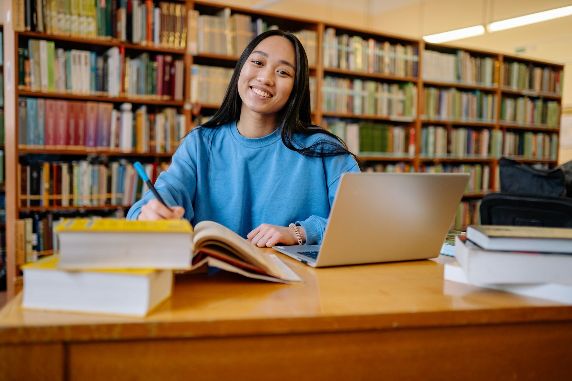 Smiling student studying in a library with books and laptop, showcasing education and focus.