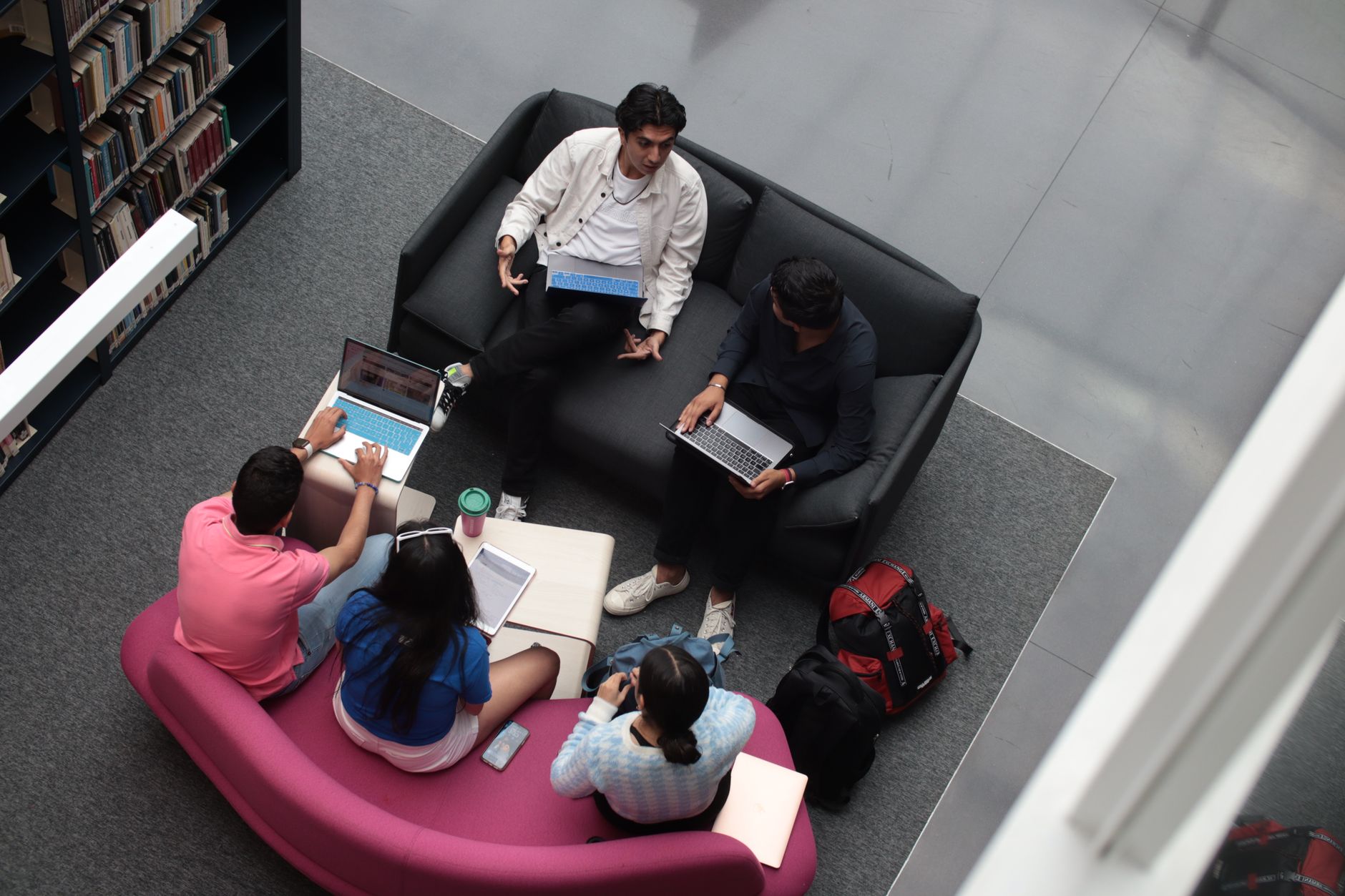 A group of diverse students using laptops and books in a library setting, studying together.