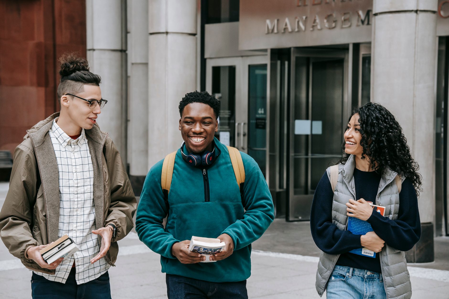 Happy young multiethnic friends in casual outfit and backpacks having conversation while walking with books in city street near building with glass doors and columns in daylight